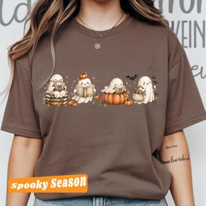 Puede incluir: Una camiseta marrón con un gráfico de cuatro fantasmas de dibujos animados. Un fantasma lleva gafas y está sentado sobre libros, otro sostiene un libro con una calabaza en la cabeza, uno está sentado sobre una calabaza leyendo y el último lleva una bolsa.