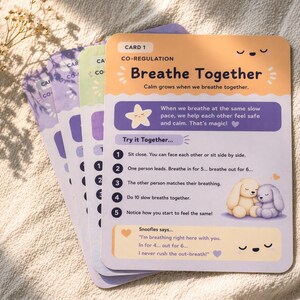 May include: A stack of colorful cards with rounded corners, featuring illustrations and text. The top card, labeled "Card 1," has the title "Breathe Together" and instructions for co-regulation. The cards have a soft, pastel color palette with cute illustrations.