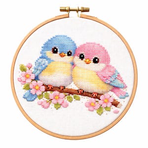 以下が含まれることがあります： ピンクの花が咲く枝に止まっている2羽の鳥のクロスステッチ刺繍。鳥は青とピンクの羽、黄色の腹、大きな黒い目を持っています。刺繍は木製のフープにセットされています。