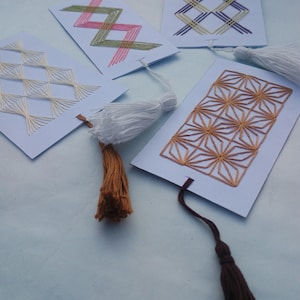 May include: Handmade bookmarks with embroidered geometric designs in various colors. Each bookmark features a different pattern, including zigzags, diamonds, and starbursts, and is finished with a tassel. The tassels are white, brown, and tan.