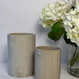 May include: Two antique stoneware crocks in a neutral color, with vertical ribbed detailing. The taller crock is on the left, and a smaller one is on the right. White flowers in a glass vase are in the background.