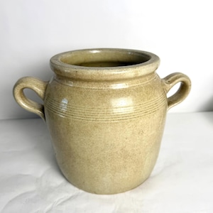 May include: A tan ceramic crock featuring a wide mouth, a thick rounded rim, and two side handles. The exterior surface has a smooth, glazed finish with subtle horizontal incised lines encircling the upper body of the vessel.
