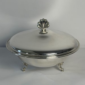 May include: A silver-colored serving dish with a lid and three ornate feet. The lid is topped with a shell-shaped handle. The dish has a beaded rim and a polished, reflective surface.