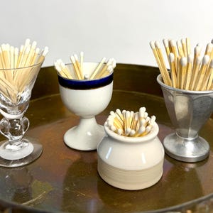 May include: Four distinct containers holding wooden matches with white tips. The collection includes a clear glass goblet, a white ceramic egg cup with a blue rim, a small white ceramic jar, and a metallic fluted cup, all arranged on a brass tray.