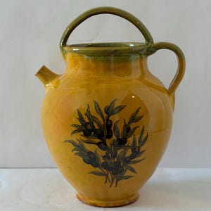 May include: A yellow ceramic pitcher with a glossy finish, featuring a dark green arched handle and a matching green rim. The body of the pitcher is decorated with a detailed black illustration of olive branches with leaves and fruit on the front.