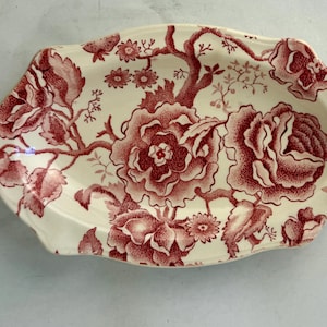 May include: An oval ceramic serving platter featuring a cream-colored base with a detailed red floral transferware pattern. The design displays large blooming roses, smaller blossoms, and leafy branches covering the entire surface of the dish.