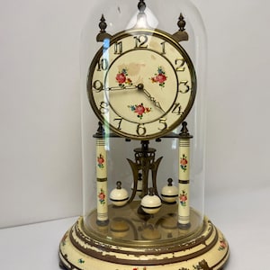 May include: An anniversary clock under a glass dome featuring a cream-colored face with gold numerals and floral motifs. The base and pillars are cream with floral accents and brown trim. The clock has a rotating pendulum with three white spheres.