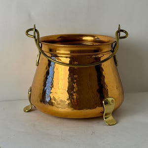 May include: A polished copper pot with a hammered texture, featuring a wide circular opening, a curved brass handle attached to two side loops, and three smooth brass feet extending from the base to support the vessel's rounded body.