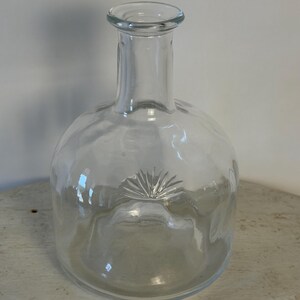 May include: Clear glass bottle with a round base and a narrow neck. The bottle has a decorative sunburst design on the front. The bottle is empty and sits on a light-colored surface.