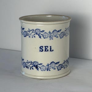 May include: A cylindrical ceramic container with an off-white finish, featuring blue floral vine patterns encircling the top and bottom rims. The word SEL is printed in blue capital letters on the center of the front side of the vessel.