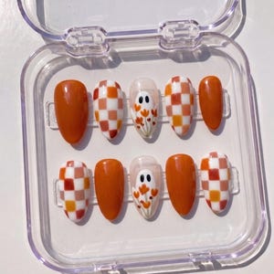 May include: A set of ten almond-shaped press-on nails in a clear plastic storage case. The designs include solid burnt orange nails, a white and orange checkered pattern, and a clear base with a white ghost figure holding orange flowers.