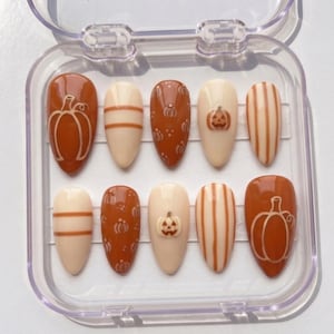 May include: A set of ten almond-shaped press-on nails in a clear plastic case. The nails feature a fall-themed design with alternating cream and burnt orange colors, decorated with hand-painted pumpkin outlines, vertical stripes, and small jack-o'-lanterns.