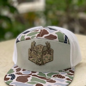 May include: A five-panel trucker hat featuring a camouflage pattern in shades of brown, green, and light blue on the brim and crown. The front panel is a solid light gray fabric with a centered, detailed patch of a deer head with antlers.