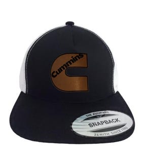 May include: Black and white snapback trucker hat with the Cummins logo in brown. The hat features a curved brim and a mesh back. The text "THE AUTHENTIC SNAPBACK ZENITH SINCE 1980" is printed on the tag.