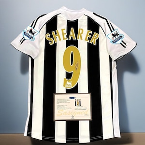 May include: A black and white striped soccer jersey with the name "SHEARER" and the number "9" in gold. The jersey has the Barclays Premier League logo on the sleeves and a certificate of authenticity.