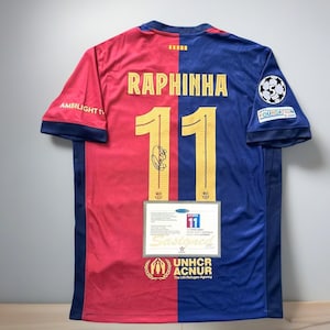 May include: A red and blue soccer jersey with the name "RAPHINHA" and the number "11" in gold. The jersey has a signature and a "Signed" certificate. The UNHCR ACNUR logo is visible.