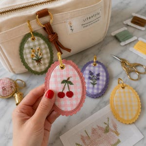 Può includere: Portachiavi ricamati di forma ovale con motivi floreali su tessuto a quadretti vichy. I portachiavi presentano motivi di ciliegie, lavanda e girasoli. Sullo sfondo una borsa color crema con la scritta "lifestyle". Sono visibili anche accessori per cucire.