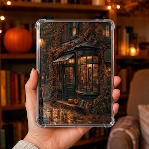 Op de afbeelding: Een doorzichtige tablethoes met een bedrukte illustratie van de buitenkant van een donkere, gezellige boekhandel in de nacht. De afbeelding toont een winkelpui met grote ramen, herfstbladeren, pompoenen op de grond en een natte, reflecterende straat.