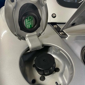 Könnte beinhalten: Nahaufnahme einer silbernen Tankklappe, die geöffnet ist, um einen schwarzen Tankdeckel freizulegen. Die Tankklappe hat einen grünen Aufkleber mit weißem Text, der "DED PREMIUM E1 SUPER SANS PLOMB" lautet. Der Tankdeckel ist schwarz und rund.