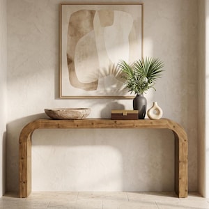 May include: A wooden console table with a curved design, holding a wooden bowl, a small box, a dark vase with greenery, and a cream-colored decorative vase. An abstract painting hangs above the table.