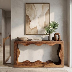 May include: A wooden console table with a wavy edge design, holding a decorative bowl, books, a vase with greenery, and a brown sculpture. A framed abstract painting hangs above the table. The room has a neutral color palette.