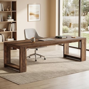 May include: A rectangular dark brown wood grain desk with a thick frame and open side supports. The desk surface holds a silver laptop, a dark mug, and a notebook. A light gray upholstered office chair with a five-point base sits behind the desk.
