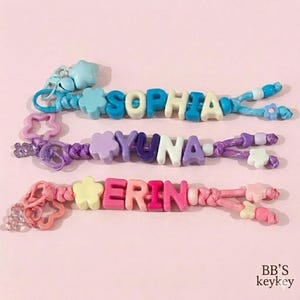 May include: Three personalized keychains with names spelled out in colorful letter beads. The names are Sophia, Yuna, and Erin. Each keychain features decorative beads and charms in shades of blue, purple, and pink, against a light pink background.