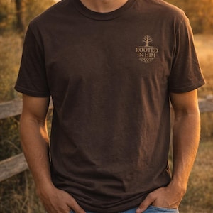 May include: Brown crew neck t-shirt with the text "ROOTED IN HIM" and a tree graphic. The shirt is made of a soft material and is paired with blue jeans. The image is taken outdoors with a natural background.