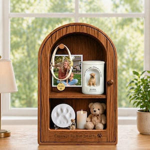 Op de afbeelding: Een houten gedenkkast voor huisdieren met een gewelfde bovenkant. Binnenin bevinden zich planken met een foto, een keramische urn met een hondenbeeldje, een voetafdruk, een kaars en een teddybeer. De kast is gegraveerd met de woorden "Forever in our hearts."