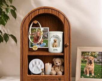 May include: An arched wooden memorial display case featuring a photo of a person with a dog, a white ceramic urn with a dog figurine, a paw print clay mold, a small glass vial, a white pillar candle, and a tan plush teddy bear inside.