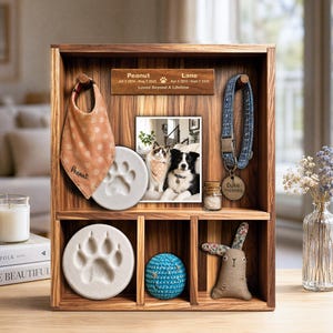 May include: A wooden shadow box display case featuring a memorial plaque with the text Peanut, Lane, dates, and Loved Beyond A Lifetime. It holds a pet bandana, a blue collar with a tag, a photo of two pets, two paw print casts, a small jar, a blue ball, and a fabric rabbit toy.
