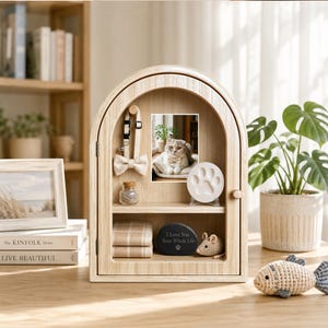 May include: A light wood arched display cabinet featuring a photo of a cat, a pet collar with a bow tie, a small glass jar, a white paw print casting, a folded plaid cloth, a black stone with the text I Love You Your Whole Life, and a toy mouse.