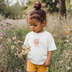 May include: A young child wearing a white t-shirt with a cartoon bee graphic, paired with yellow shorts. The child is holding a bouquet of white and yellow flowers in a field of tall grass and wildflowers.