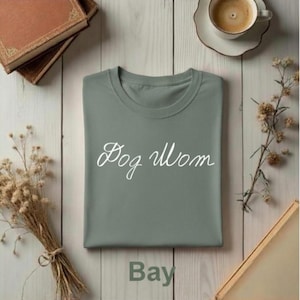 May include: A sage green t-shirt with the words "Dog Mom" in white script. The word "Bay" is printed in a sans-serif font at the bottom of the shirt. The shirt is folded neatly on a light wood surface.