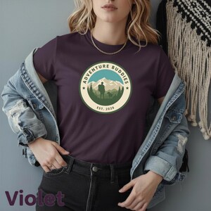 Puede incluir: Camiseta violeta con un gráfico circular que dice "Adventure Buddies" alrededor de una escena de montaña con una persona y un perro. El texto "Est. 2026" está debajo. La camiseta se usa con una chaqueta vaquera.