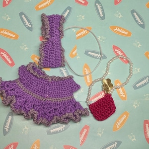 Op de afbeelding: Een handgemaakt gehaakt poppenoutfit in paarse en grijze tinten, inclusief rok, top en polsbandjes. Een parelketting met een rode handtas en een zilveren haaraccessoire zijn ook inbegrepen. De achtergrond heeft een lichtblauw patroon met surfplankafbeeldingen.