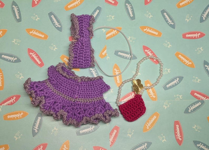 Op de afbeelding: Een handgemaakt gehaakt poppenoutfit in paarse en grijze tinten, inclusief rok, top en polsbandjes. Een parelketting met een rode handtas en een zilveren haaraccessoire zijn ook inbegrepen. De achtergrond heeft een lichtblauw patroon met surfplankafbeeldingen.