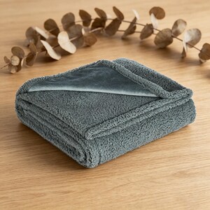 May include: A folded, gray plush blanket with a soft, textured surface. The blanket is folded neatly, revealing a contrasting smooth, gray interior. The blanket is on a wooden surface, with dried leaves in the background.
