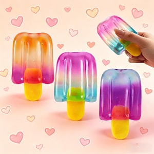 Könnte beinhalten: Drei bunte Spielzeuge in Form von Eis am Stiel in den Farbtönen Orange, Rosa, Blau und Lila. Jedes hat einen gelben Stiel und einen Farbverlauf. Eines wird von einer Hand gehalten. Der Hintergrund zeigt rosa und gelbe Herzen.