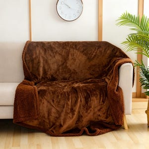 May include: A brown, plush throw blanket draped over a beige sofa. The blanket has a soft, textured appearance and is folded to display the sherpa lining. A clock and a potted plant are visible in the background.