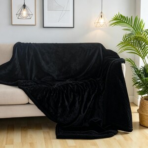 May include: A black plush throw blanket draped over a beige sofa. The blanket has a soft, textured appearance and covers the entire sofa. A houseplant in a woven basket sits to the right. Decorative artwork and lighting fixtures are visible in the background.