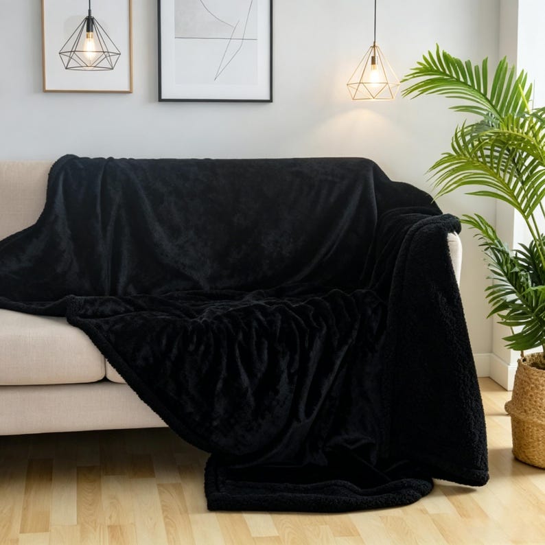 May include: A black plush throw blanket draped over a beige sofa. The blanket has a soft, textured appearance and covers the entire sofa. A houseplant in a woven basket sits to the right. Decorative artwork and lighting fixtures are visible in the background.