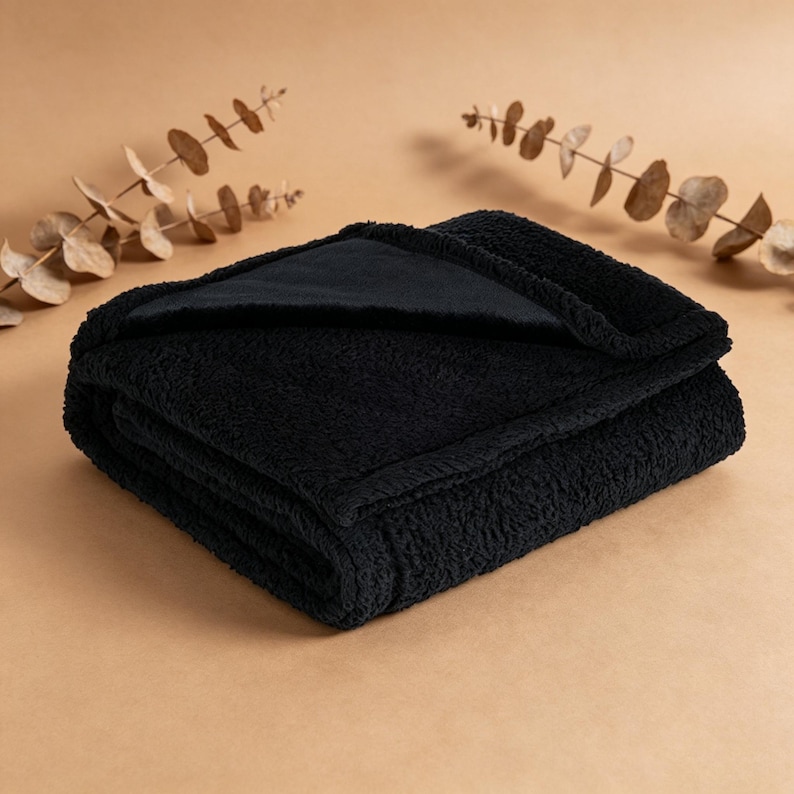 May include: A folded black plush blanket with a soft, textured surface. The blanket is made of a thick, cozy material, perfect for snuggling. The blanket is folded neatly, with a contrasting black fabric visible on the inside.