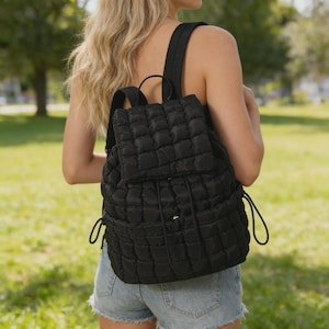 May include: A black quilted backpack featuring a square-patterned texture, a top flap with a drawstring closure, and adjustable shoulder straps. The bag has a structured design with side pockets and a main compartment secured by a flap and cord.
