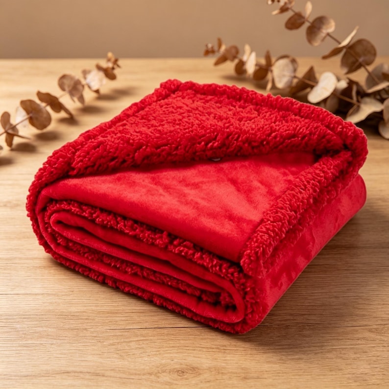 May include: A folded red blanket with a plush, textured border and a smooth, velvety interior. The blanket is neatly stacked, showcasing its layered construction. The warm red color suggests comfort and coziness. The blanket is on a wooden surface.