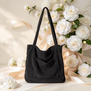 May include: A black corduroy tote bag featuring a vertical ribbed texture, two long shoulder straps, and a single open front pocket. The bag has a rectangular shape with a zippered top closure and is constructed from a soft, textured fabric.