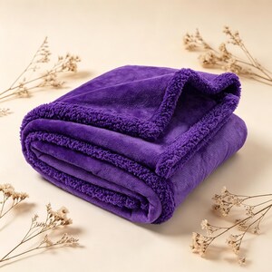 May include: A folded, plush purple blanket with a sherpa trim. The blanket is a deep violet color and appears soft and cozy. The sherpa trim adds texture and warmth to the blanket. The blanket is folded neatly.