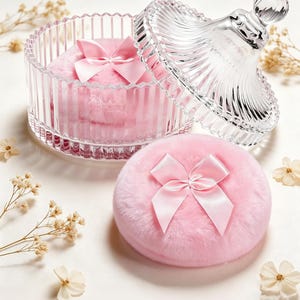 May include: A clear glass jar with a lid, containing pink powder puffs with satin bows. One puff rests outside the jar. The jar and lid have a ribbed design. Decorative flowers are scattered around the objects.
