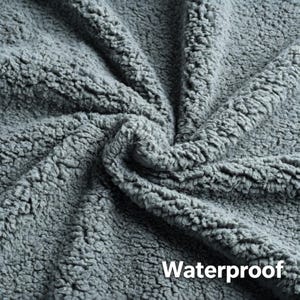 May include: Close-up of a gray, textured fabric with a plush, sherpa-like appearance. The word "Waterproof" is printed in white on the lower right corner. The fabric is folded, showing the depth and texture of the material.