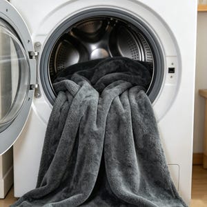 May include: A gray plush blanket partially inside a white washing machine. The blanket is a soft, textured material, with a darker gray color inside the machine. The washing machine door is open, revealing the interior.